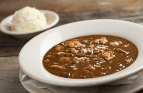 A bowl of gumbo with shrimp and rice, served alongside a small dish of white rice on a wooden table.