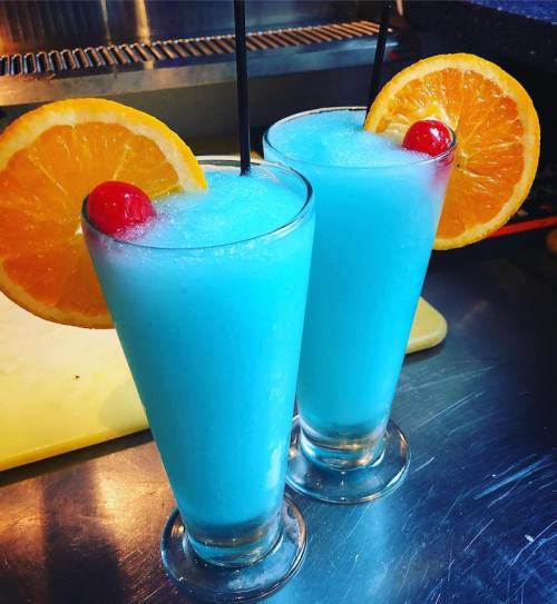 Two blue cocktails in tall glasses, garnished with orange slices and cherries, on a stainless steel surface.