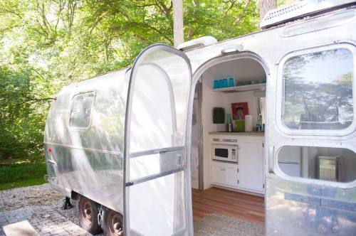 A vintage silver trailer with an open door, revealing a cozy interior with a kitchenette and greenery.