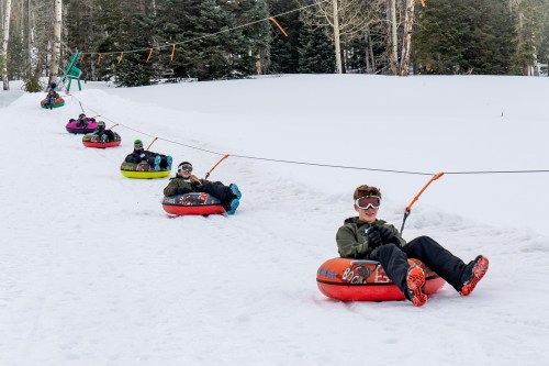 A group of people tubing down a snowy hill, connected by a tow line, surrounded by trees.
