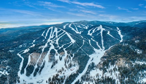 Aerial view of snow-covered mountains with ski trails and trees under a clear blue sky.
