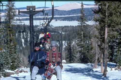 Skiers on a chairlift surrounded by snow-covered trees and mountains in the background.
