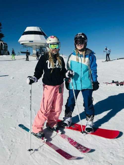 Two young girls in ski gear pose on a snowy slope, with a ski lift and other skiers in the background.