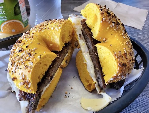A split yellow bagel filled with cream cheese and a meat patty, sprinkled with sesame seeds, on a tray.