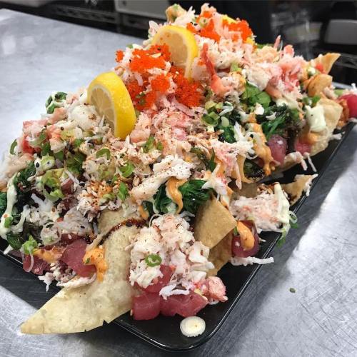 A large platter of nachos topped with crab, tuna, green onions, spicy mayo, and lemon slices, garnished with orange roe.