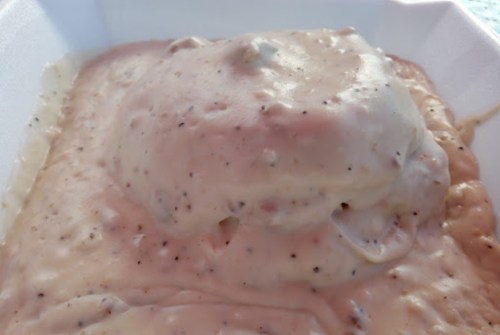 A close-up of a creamy, beige sauce with specks, covering a mound of food in a white takeout container.