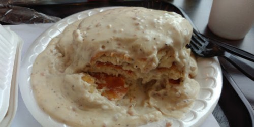 A plate of biscuits smothered in creamy sausage gravy, partially eaten, on a black tray with utensils.