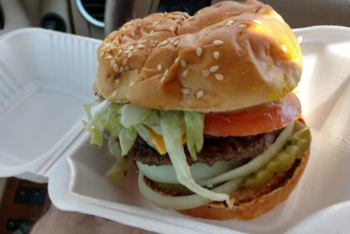 A sesame seed bun burger with lettuce, tomato, onion, pickles, and a beef patty, served in a takeout container.