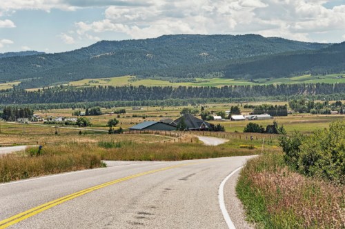 A winding road leads through a lush green landscape with mountains in the background and farms dotting the valley.