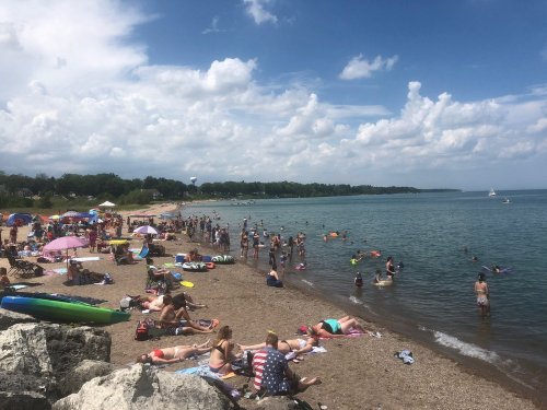 A crowded beach scene with people sunbathing, swimming, and colorful umbrellas under a partly cloudy sky.