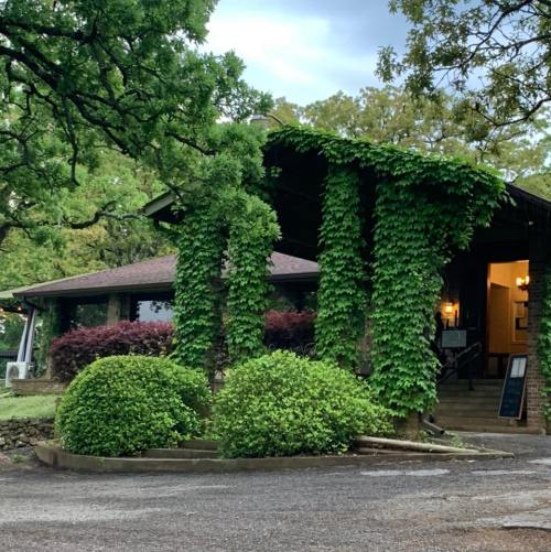 A charming building covered in green vines, surrounded by lush bushes and trees, set against a cloudy sky.