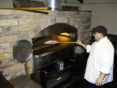 A chef in a white coat uses a peel to place dough into a stone oven.