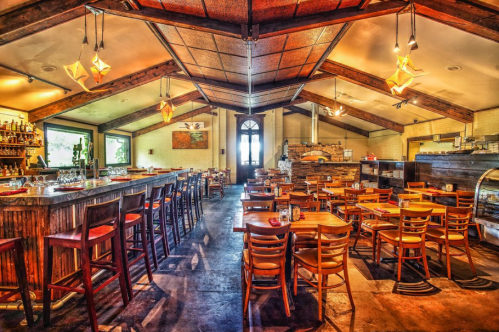 A spacious restaurant interior with wooden beams, a bar, and tables arranged for dining. Warm lighting creates a cozy atmosphere.
