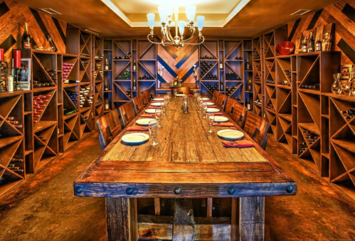 A rustic dining room with a long wooden table set for a meal, surrounded by wine racks and warm lighting.