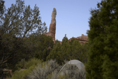 A tall rock formation rises above green shrubs and trees, with a tent set up in the foreground.