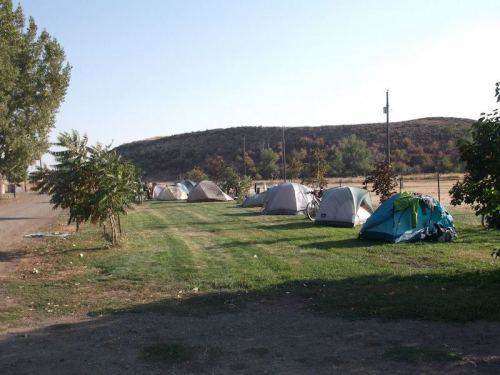 A campsite with several tents set up on grassy land, surrounded by trees and a hillside in the background.