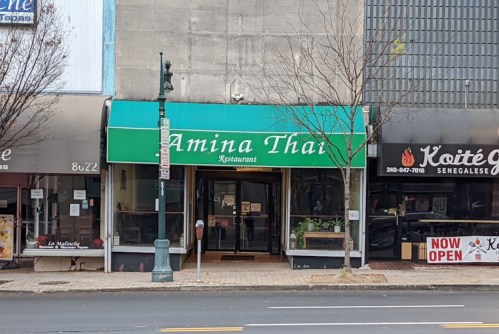Exterior of Amina Thai restaurant with a green awning, located between two other eateries on a city street.