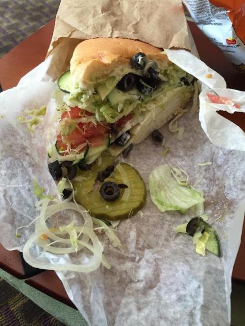 A partially unwrapped sandwich with lettuce, tomato, cucumber, olives, and pickles on a table, surrounded by crumpled paper.