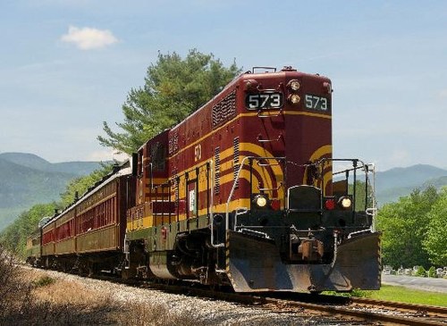 A vintage train with a red and yellow design travels along a scenic route with mountains in the background.
