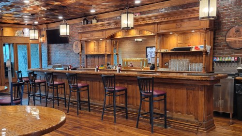 A wooden bar with high stools, brick walls, and pendant lights in a cozy pub setting.