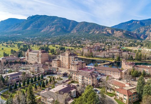 Aerial view of a resort surrounded by mountains and greenery, with a lake and buildings in the foreground.
