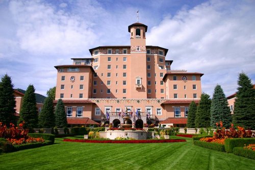A grand hotel with a pink facade, surrounded by lush green lawns and colorful flower beds under a blue sky.