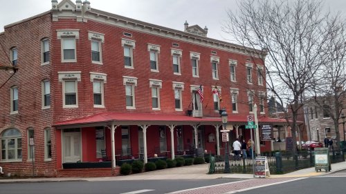 Historic red brick building with a porch, flags, and people outside, set in a small town.