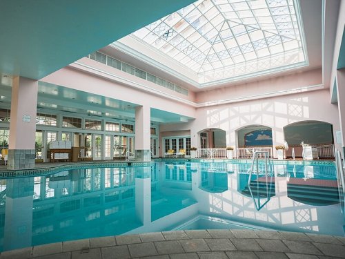 Indoor swimming pool with a glass ceiling, surrounded by light pink walls and large windows.