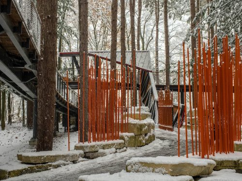 A snowy forest scene featuring orange vertical structures and a pathway, with a bridge above among the trees.