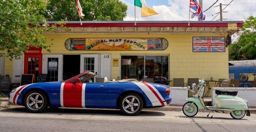 A colorful convertible car parked outside a quirky shop with flags and a vintage scooter nearby.