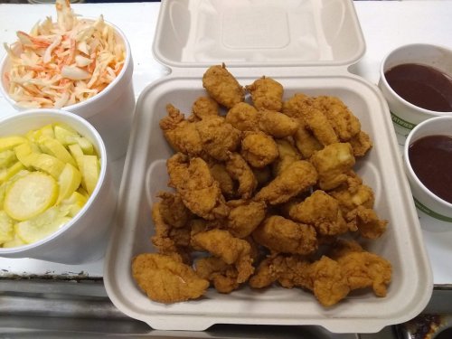 A takeout container filled with fried chicken, served with coleslaw, pickles, and two cups of sauce.
