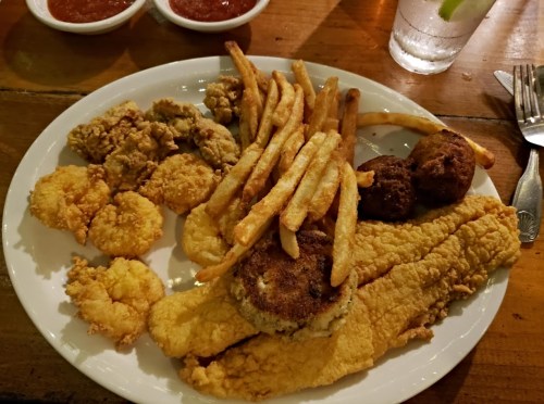 A plate of fried fish, shrimp, chicken, and French fries, served with dipping sauces on the side.