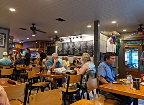 A busy restaurant interior with diners at wooden tables, a chalkboard menu, and a casual atmosphere.