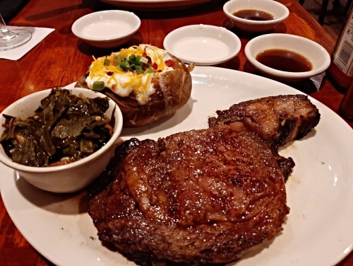 A plate featuring a juicy steak, a loaded baked potato, and a side of collard greens, with dipping sauces in the background.