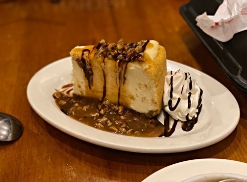 A slice of cheesecake topped with nuts and chocolate drizzle, served with a side of whipped cream on a plate.