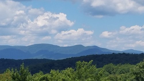 A scenic view of rolling mountains under a blue sky with fluffy white clouds and green trees in the foreground.