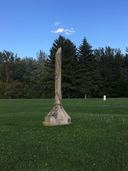 A tall, weathered monument stands in a grassy field, surrounded by trees under a clear blue sky.