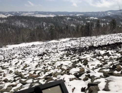 A snowy landscape with scattered rocks, rolling hills, and distant mountains under a partly cloudy sky.