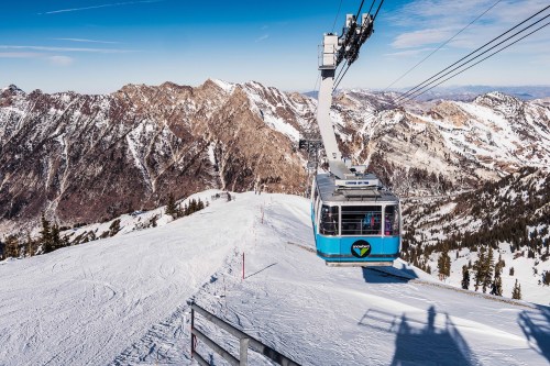 A ski gondola ascends over snow-covered mountains under a clear blue sky.