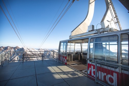 A cable car station with panoramic mountain views under a clear blue sky. Snow-capped peaks in the background.