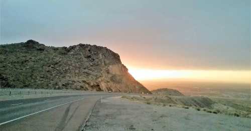 A winding road curves around a rocky hill at sunset, with a warm glow illuminating the horizon.