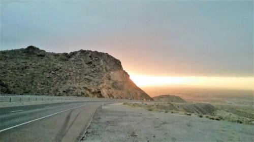 A winding road curves around a rocky hill at sunset, with a warm glow illuminating the horizon.