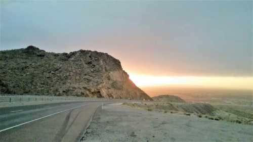 A winding road curves around a rocky hill at sunset, with a warm glow illuminating the horizon.