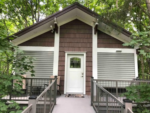 A cozy cabin with a brown and gray exterior, surrounded by trees, featuring a front porch and a white door.