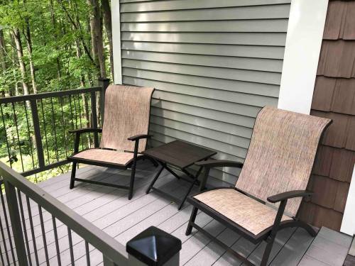 Two outdoor chairs and a small table on a balcony, surrounded by green trees and a wooden house exterior.