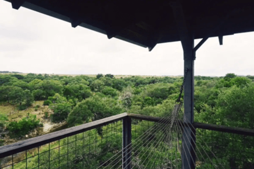 View from a wooden deck overlooking a lush green landscape with trees and a cloudy sky. A hammock is visible.