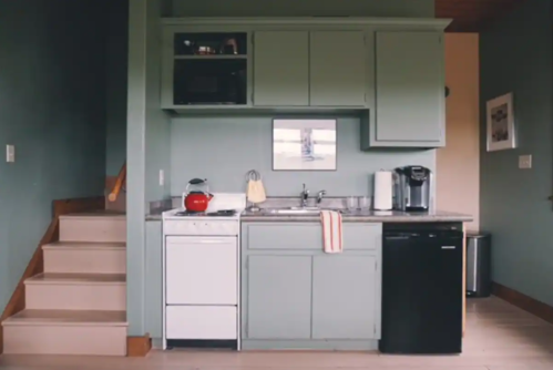 A modern kitchen with green cabinets, a sink, and appliances, next to a staircase in a cozy setting.