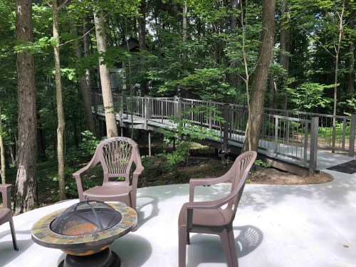 A serene outdoor space with a fire pit and chairs, surrounded by trees and a wooden walkway in the background.