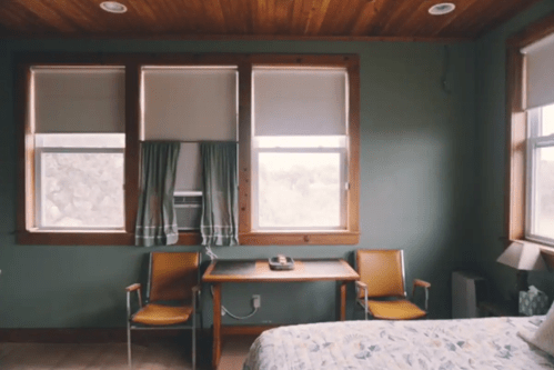 A cozy room with two windows, a bed, a desk with a phone, and two chairs, featuring wooden walls and green paint.