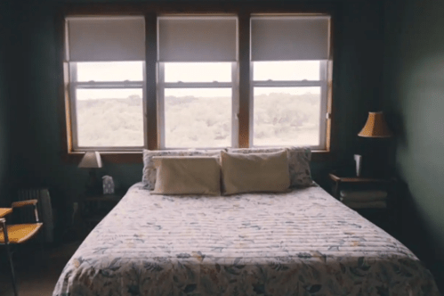Cozy bedroom with a floral bedspread, two pillows, and large windows overlooking greenery.